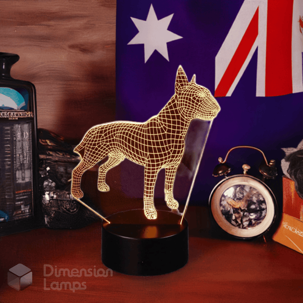 A warmly lit 3D Bull Terrier lamp with a wireframe structure, displayed on a wooden surface alongside an Australian flag and a classic alarm clock. A decorative gift for Bull Terrier owners and dog lovers in Australia. A dark bottle is also visible in the background.