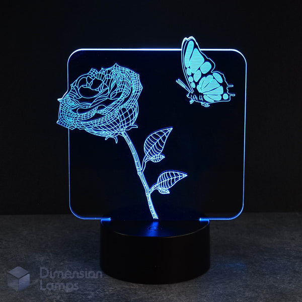 A cyan LED light forms the shape of a rose flower on a stem with leaves and a butterfly in this 3D lamp, set against a dark backdrop. This floral and insect lamp offers a unique and eye-catching lighting option.