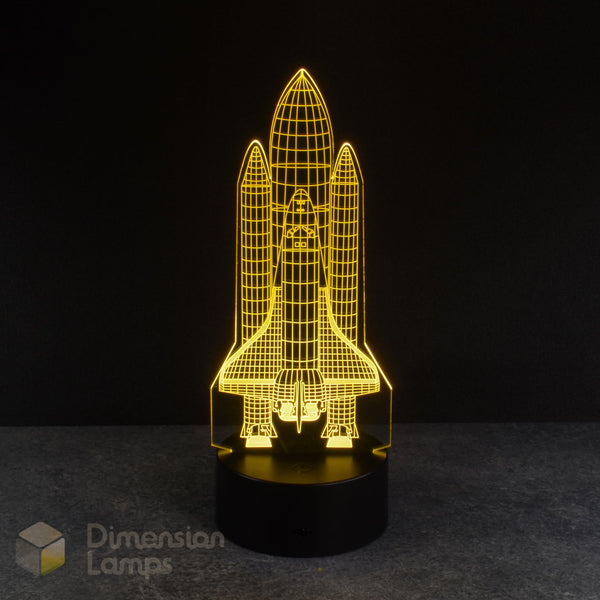 A yellow 3D lamp displays an etched design of a space shuttle, illuminated on a black base against a dark backdrop. An awesome nightlight for boys rooms across Australia.