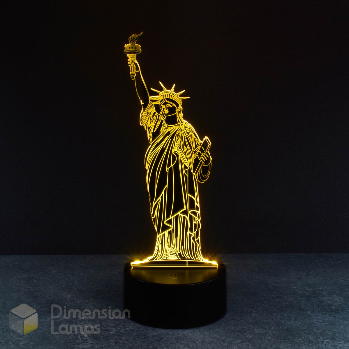 A 3D illusion lamp showcases the silhouette of the Statue of Liberty, a well-known American icon, illuminated in yellow and emanating upwards from a black base against a dark background. An engaging and distinctive travel souvenir.
