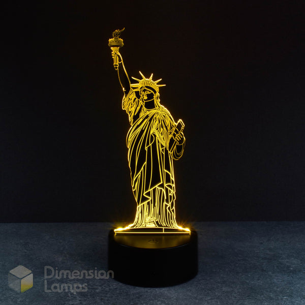 A 3D illusion lamp showcases the silhouette of the Statue of Liberty, a well-known American icon, illuminated in yellow and emanating upwards from a black base against a dark background. An engaging and distinctive travel souvenir.