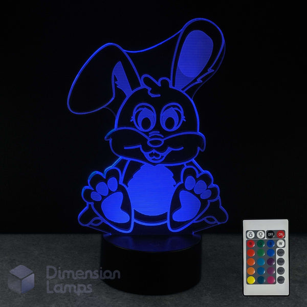 A 3D optical illusion lamp of a cartoon bunny, glowing with blue light on a black base, shown with its remote control against a black background.