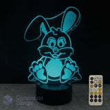 A 3D optical illusion LED lamp of a cartoon bunny glowing bright cyan n a black base. A 7 colour remote control and the Dimension Lamps watermark.