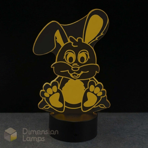 A 3D acrylic LED night light shaped like a cartoon bunny, featuring a yellow design on a black cylindrical base, set against a dark background.