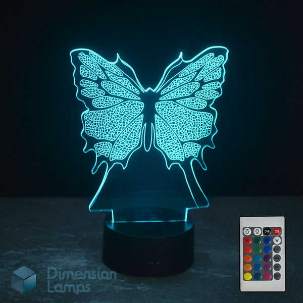 A 3D optical illusion LED night light shaped like a butterfly, glowing with a cyan light, shown on a dark surface with its remote control in the bottom right corner. Butterfly lamp gift. 