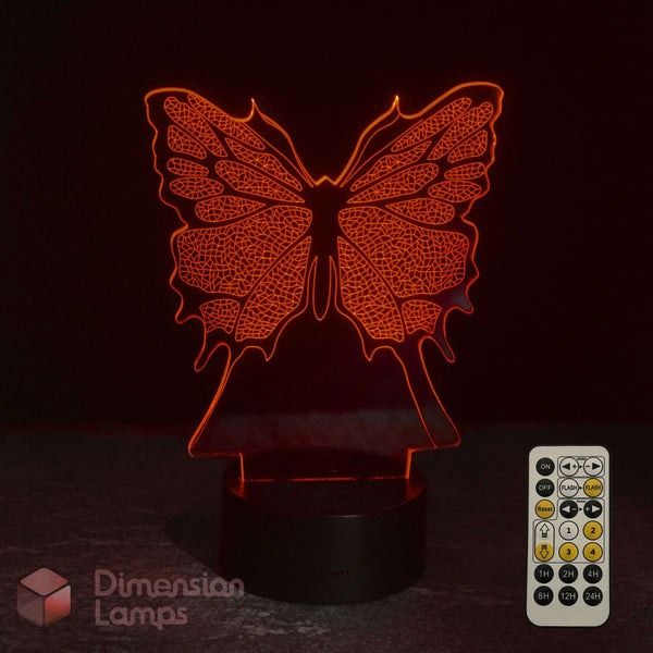 A 3D optical illusion LED night light shaped like a butterfly, glowing with a red light on a dark surface with its 7 colour sleep timer remote control in the bottom right corner. Girls butterfly lamp. 