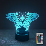 A butterfly-shaped 3D optical illusion night light glowing with a bright cyan-blue light, set on a black base with a 16 colour remote control shown in the bottom right corner. The background is a very dark, near-black studio setting.