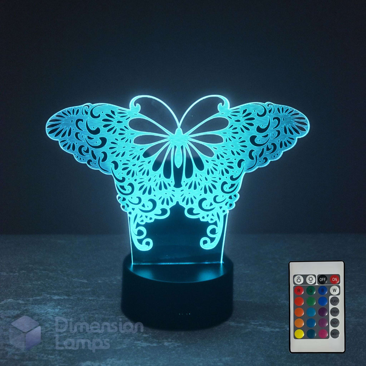 A butterfly-shaped 3D optical illusion night light glowing with a bright cyan-blue light, set on a black base with a 16 colour remote control shown in the bottom right corner. The background is a very dark, near-black studio setting.