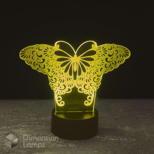A butterfly-shaped 3D optical illusion night light glowing with a warm yellow light, set against a dark background on a gray surface. A 'Dimension Lamps' watermark is visible in the bottom-left corner.