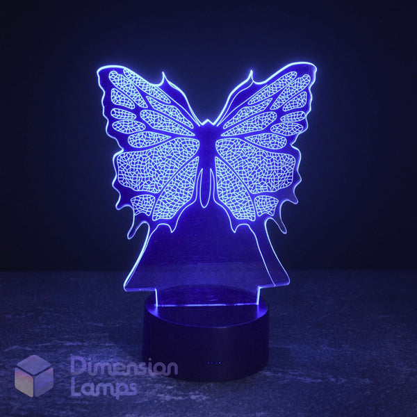 A butterfly-shaped LED night light glowing with a blue-purple light on a dark base, set against a dark background. A 'Dimension Lamps' logo watermark is visible in the bottom-left corner.