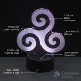 A 3D optical illusion lamp with a glowing white triskelion design on a black base, featuring text callouts detailing its features like '7 colour modes', 'precision laser etching', and 'clear acrylic display'. Celtic art gift.