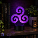 A purple Celtic triskelion night light with a glowing geometric design on a black base, displayed on a wooden shelf next to books, a plant, and a candle. Celtic art lovers gift idea.