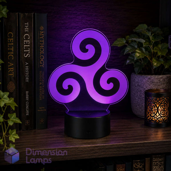 A purple Celtic triskelion night light with a glowing geometric design on a black base, displayed on a wooden shelf next to books, a plant, and a candle. Celtic art lovers gift idea.