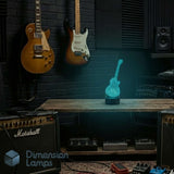 A glowing blue guitar-shaped night light displayed on a wooden table in a music studio setting, with electric guitars, Marshall amplifiers, and audio equipment in the background.