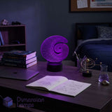 A glowing purple Fibonacci spiral 3D LED lamp sits on a study desk in a dimly lit bedroom with books and a sketchbook. Design student gift idea. 