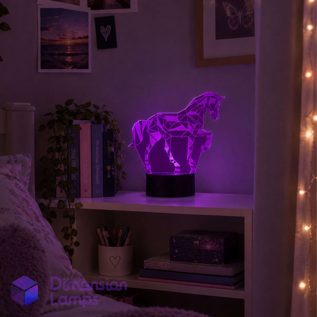 A purple geometric unicorn lamp on a black base, displayed on a white shelf in a cosy bedroom setting with warm string lights and framed wall art.