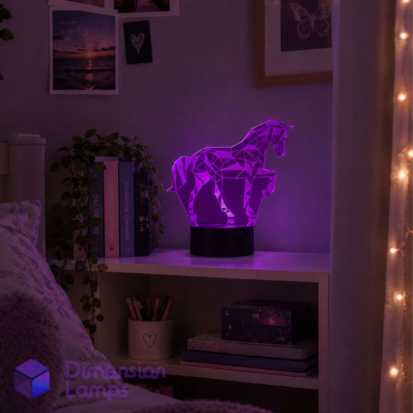 A purple geometric unicorn lamp on a black base, displayed on a white shelf in a cosy bedroom setting with warm string lights and framed wall art.