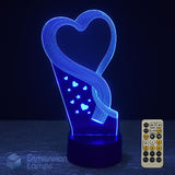 Blue 3D optical illusion LED night light shaped like a heart with a swirling ribbon design and small heart and star accents, shown on a dark surface with a small remote control beside it.