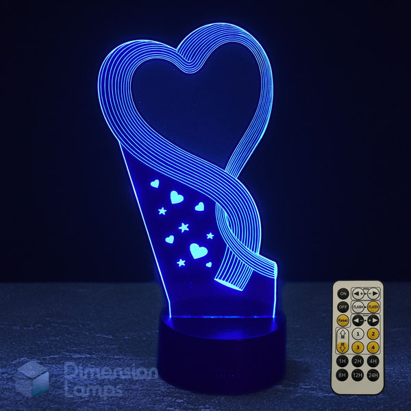 Blue 3D optical illusion LED night light shaped like a heart with a swirling ribbon design and small heart and star accents, shown on a dark surface with a small remote control beside it.