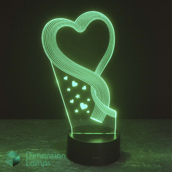 A 3D optical illusion LED lamp by Dimension Lamps, featuring a glowing green heart and ribbon design on a black base, set against a dark background.