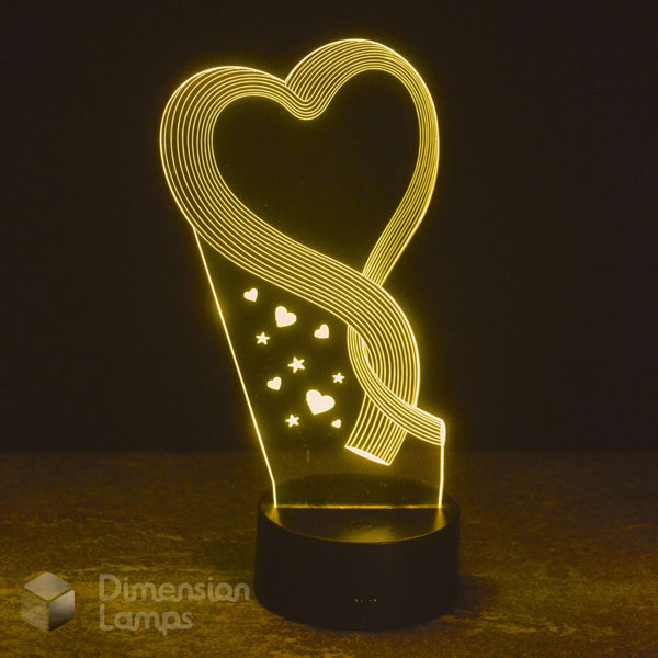 A glowing LED acrylic lamp shaped like a heart with a ribbon-like swirl and small heart and star cutouts, sitting on a black base against a dark background. A 'Dimension Lamps' watermark is visible in the lower left.