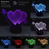 A 3D illusion LED lamp shaped like two interlocking hearts with arrows, shown in multiple color modes on a dark background. The left large panel shows the lamp lit in purple, with smaller panels showing other available colors: cyan, white, blue, red, green, and yellow.