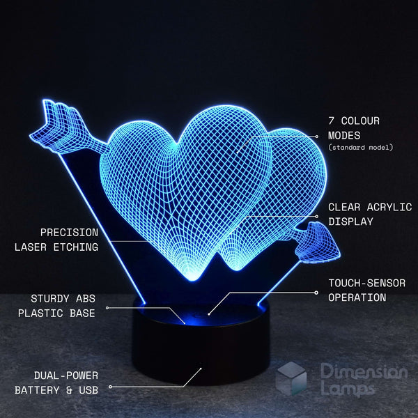 A 3D illusion LED lamp shaped like two interlocking hearts pierced by arrows, glowing in blue light on a black base. White text callouts highlight features such as 7 colour modes, clear acrylic display, precision laser etching, sturdy ABS plastic base, touch-sensor operation, dual-power battery & USB, and the Dimension Lamps brand.