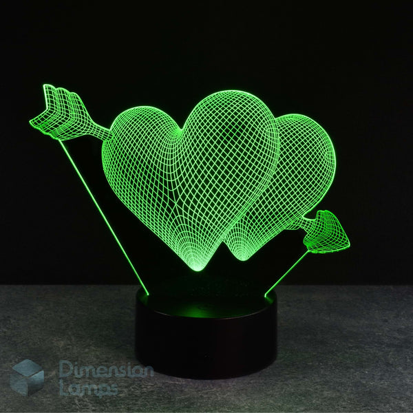 A 3D optical illusion LED night light featuring two glowing green wireframe hearts pierced by arrows, mounted on a black cylindrical base against a dark background.
