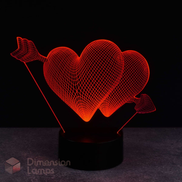 A 3D optical illusion LED night light featuring two glowing red hearts pierced by an arrow, mounted on a black cylindrical base against a dark background.