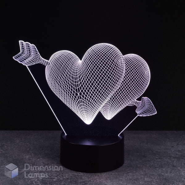 A 3D optical illusion LED night light featuring two interlocking glowing wireframe hearts pierced by an arrow, mounted on a black cylindrical base against a dark background.