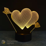 A 3D optical illusion lamp featuring two glowing wireframe hearts pierced by arrows, mounted on a black cylindrical base against a dark background. A 'Dimension Lamps' logo watermark is visible in the bottom-left corner.