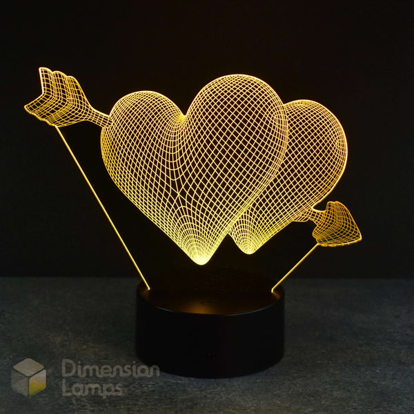 A 3D optical illusion lamp featuring two glowing wireframe hearts pierced by arrows, mounted on a black cylindrical base against a dark background. A 'Dimension Lamps' logo watermark is visible in the bottom-left corner.
