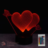 A 3D optical illusion lamp featuring two glowing red hearts pierced by arrows, mounted on a black circular base. A small remote control is shown in the lower right corner against a dark background.