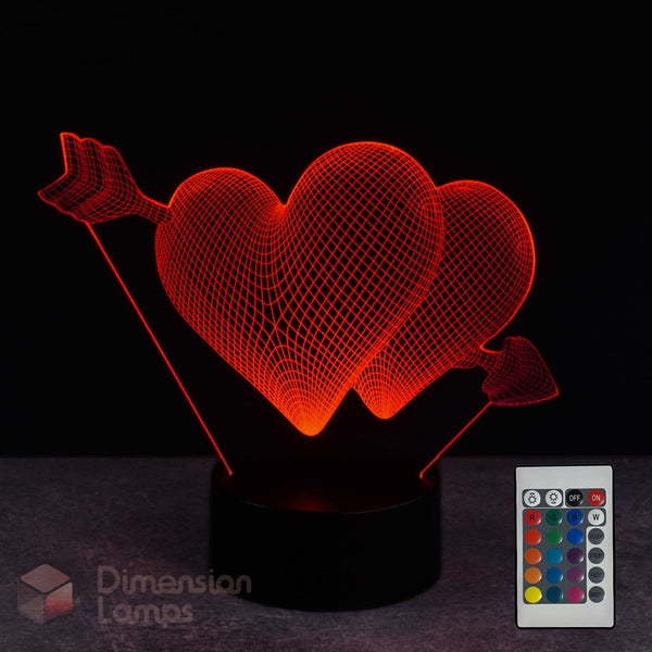 A 3D optical illusion lamp featuring two glowing red hearts pierced by arrows, mounted on a black circular base. A small remote control is shown in the lower right corner against a dark background.