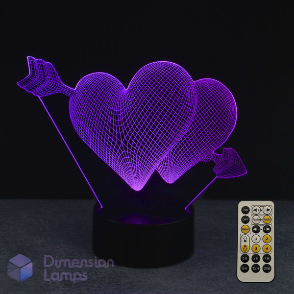 A 3D optical illusion LED night light featuring two interlocking hearts pierced by an arrow, glowing with purple light on a black cylindrical base. A white remote control is shown in the bottom right corner against a dark background.