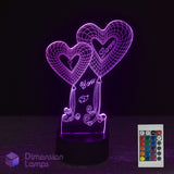 A 3D optical illusion LED night light featuring two heart-shaped balloons with 'I Love You' written on them, glowing in purple on a black base. A white remote control is shown in the bottom right corner, and a 'Dimension Lamps' logo is visible in the bottom left.