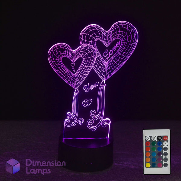 A 3D optical illusion LED night light featuring two heart-shaped balloons with 'I Love You' written on them, glowing in purple on a black base. A white remote control is shown in the bottom right corner, and a 'Dimension Lamps' logo is visible in the bottom left.