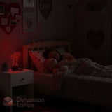 A child's bedroom scene in very low light showing a bed with a child tucked in, a bedside table with a glowing heart-shaped lamp, and heart-themed wall decorations. The watermark 'Dimension Lamps' appears in the lower left.