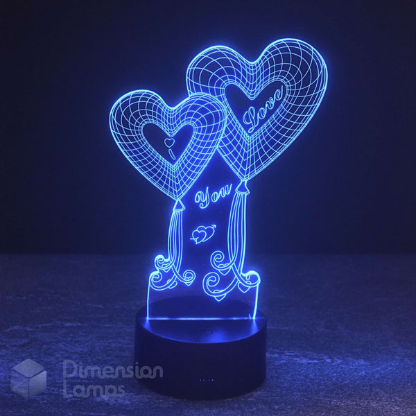 A 3D optical illusion night light by Dimension Lamps featuring two heart-shaped balloons with 'I Love You'  written inside, glowing with a blue LED light on a black base.