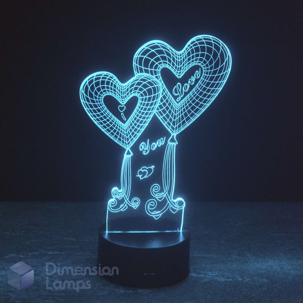 A 3D optical illusion night light by Dimension Lamps featuring two glowing heart-shaped balloons with 'I Love You' written on them, floating above a black cylindrical base against a dark background.