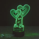 A 3D optical illusion LED night light shaped like two heart balloons with 'I Love You' written on them, glowing green on a black base against a dark background. A 'Dimension Lamps' watermark is visible in the lower-left corner.
