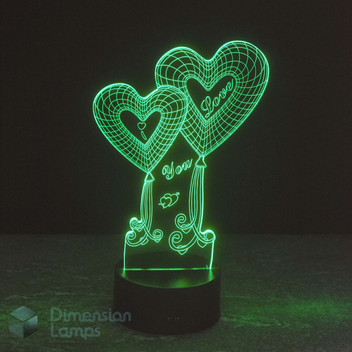 A 3D optical illusion LED night light shaped like two heart balloons with 'I Love You' written on them, glowing green on a black base against a dark background. A 'Dimension Lamps' watermark is visible in the lower-left corner.