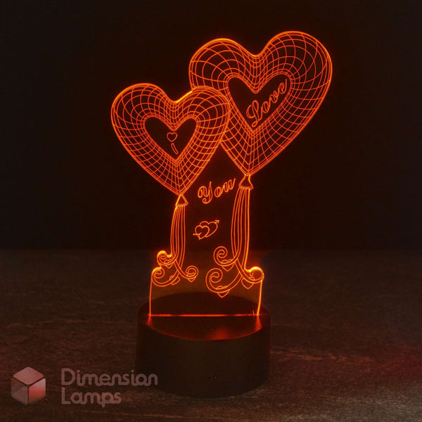 An LED acrylic 3D optical illusion lamp featuring two interlocking heart-shaped balloons with 'I Love You' written inside, glowing with a warm orange light on a black base.