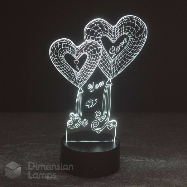 A 3D acrylic illusion night light featuring two heart-shaped balloons with 'I Love You' written inside, glowing with a white light on a black cylindrical base. The brand name 'Dimension Lamps' is visible as a watermark in the bottom-left corner.
