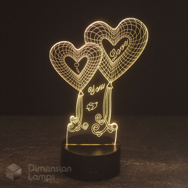 A 3D optical illusion LED night light featuring two glowing heart-shaped balloons with 'I Love You'  written on them, mounted on a black cylindrical base against a dark background.
