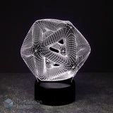 A 3D optical illusion lamp featuring a white wireframe icosahedron geometric design that creates a swirling, abstract effect, resting on a black cylindrical base against a dark background.