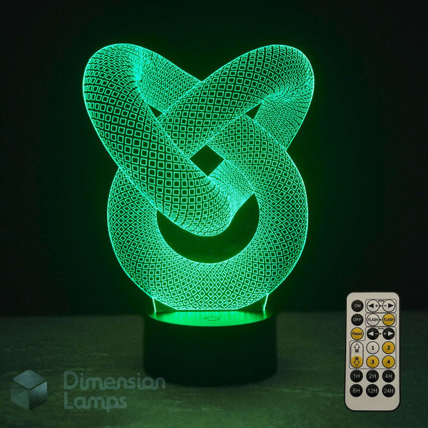 A 3D optical illusion lamp with an interlocking knot design glowing in vibrant green, shown with its remote control on a dark surface against a black background. A 'Dimension Lamps' watermark is visible in the bottom-left corner.