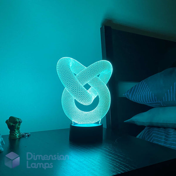 A 3D optical illusion LED night light shaped like an interwoven knot, glowing with a cyan-blue light on a dark bedside table next to a bed.