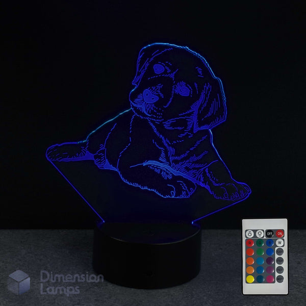 A 3D optical illusion night light shaped like a puppy, glowing with blue light on a black base, shown alongside a 16 colour remote control and a 'Dimension Lamps' logo watermark.