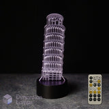 An LED 3D optical illusion night light shaped like the Leaning Tower of Pisa, glowing with a purple light on a black base, shown with the 7 colour remote control against a dark background.